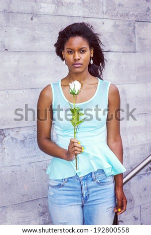 Girl Missing you. Wearing a green tank top, drop earrings, blue jeans, holding a white rose, a young woman is standing against the wall outside in wind, waiting for you. Instagram Hudson effect.