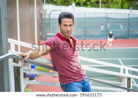 Man waiting for you. Wearing a red Polo shirt, blue jeans, stretching arms on the railing, a handsome, sexy, middle age guy is standing by a tennis court, passionately looking at you.