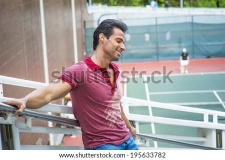 Man thinking of you. Wearing a red Polo shirt, blue jeans, stretching arms on the railing, a handsome, sexy, middle age guy is standing by a tennis court, smiling, thinking.