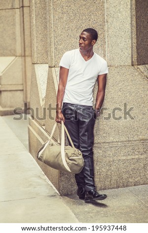 Street Fashion. Wearing a white V neck T shirt, pants, leather shoes, carrying a bag,  a young black college student is standing against a column outside an office building,  looking away.