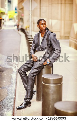 Young College Student. Carrying a bag, holding a laptop computer, a young handsome black guy is sitting on street, relaxing, waiting, thinking.
