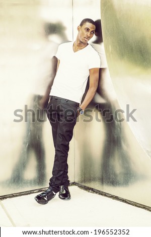 Reflection. Wearing a white V neck T shirt, pants, leather shoes, wristwatch, hands in pocket,  a young black handsome man is leaning back against  metal mirror walls, lost in thought.