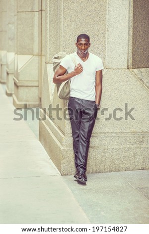 Street Fashion. Wearing a white V neck T shirt, pants, leather shoes, carrying a shoulder bag,  a young black college student is standing outside an office building, looking at you.