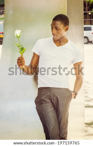 Falling in love.  Wearing a white V neck T shirt, a hand holding a white rose, a young black guy is leaning back against  a metal structure on street, passionately looking at, smiling, thinking.
