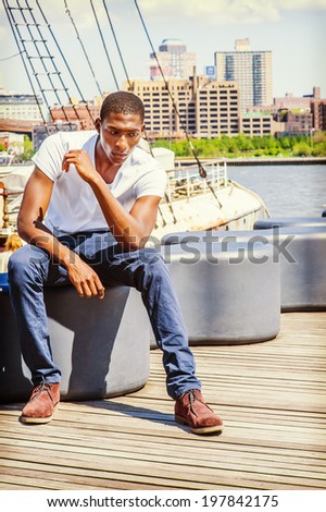 Man Thinking Outside. Wearing a white T shirt, blue pants, brown boot shoes, a young black guy is sitting on deck, looking down, relaxing, lost in thought. The background is a harbor.