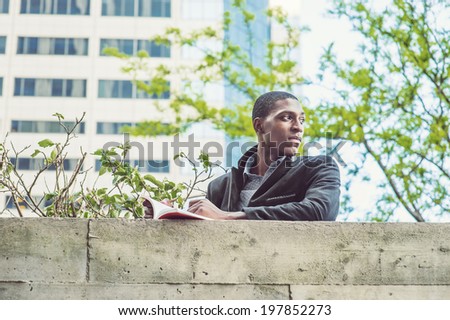 Young black man reading outside. A young black college student is standing by the top of the wall outside a building, reading a book, looking around, waiting for you.