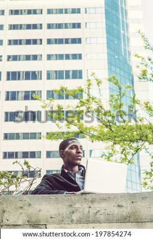 Young black man working outside. A young black college student is standing by the top of the wall outside an office building,  working on a computer.