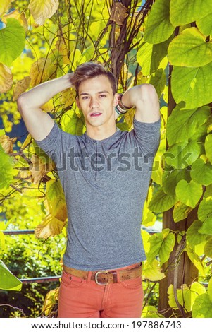 Man Casual Fashion. Dressing in a gray long sleeves with roll-tab Henley shirt, red jeans, hands putting in the back of head, a young handsome guy is standing by plants with big leaves, looking at you