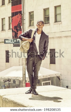 Street Fashion. Wearing a white under wear, fashionable jacket, pants, leather shoes, carrying a shoulder bag,  a young black college student is standing on Wall Street, hopefully looking at you.