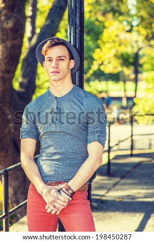 Man Thinking Outside. Dressing in a gray long sleeves with roll-tab Henley shirt, red jeans, wearing a woolen Fedora hat, a young handsome guy is leaning on a black pole, tired, lost in thought.