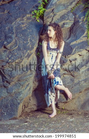 Teen Fashion. Dressing in patterned long dress, chunky chain bracelet, arm cuff bracelet, a teenager girl with curly long hair is standing against rocky wall, looking around, relaxing, waiting for you