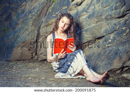 Girl Reading Outside. Wearing long dress, bracelet, barefoot, a pretty teenage college student with curly long hair is sitting on ground against rocks, holding a red book, reading, relaxing.