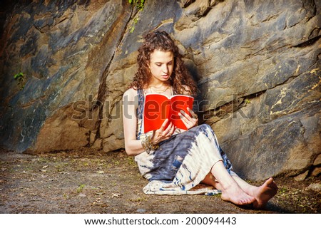 Girl Reading Outside. Wearing long dress, bracelet, barefoot, a pretty teenage college student with curly long hair is sitting on ground against rocks, holding a red book, reading, thinking, relaxing.