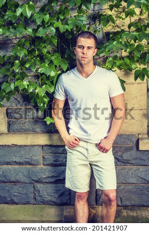 Portrait of young man. Wearing a white V neck T shirt, light yellow shorts, a young guy is standing against a wall with green ivy leaves, confidently looking at you.
