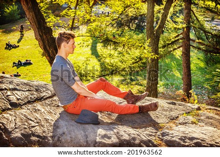 Lonely Man. Dressing in a gray long sleeves with roll-tab Henley shirt, red jeans, brown boot shoes, a Fedora hat on side, a young guy is sitting on rocks in Autumn, relaxing, thinking.