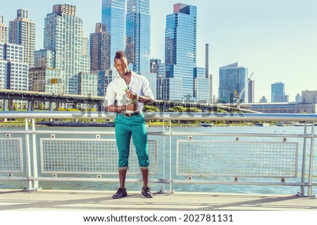 Missing You. Wearing white shirt, green pants, leather shoes, a young black guy with mohawk hair is standing in the front of buildings, holding white rose, looking down, thinking. Urban Casual Fashion