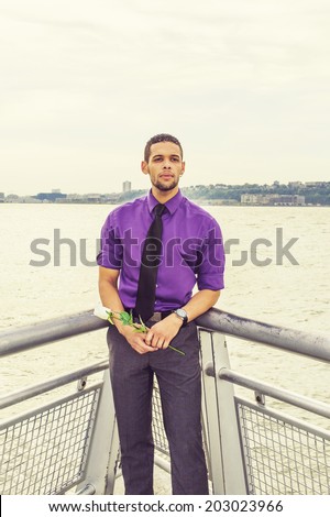 Young Man Waiting for You. Dressing in a purple shirt, gray pants, a black neck tie, holding a white flower, a young guy with beard, mustache is waiting for you by the river.