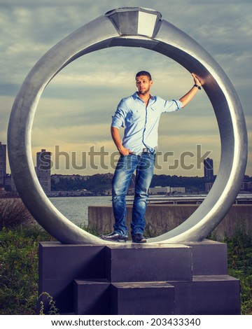 Man Missing You. Wearing a light blue shirt, blue jeans, leather shoes, a young handsome guy with a little beard, mustache is standing by a ring structure, waiting for you, symbolic for engagement.