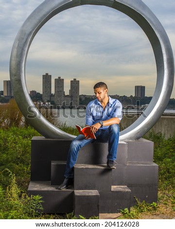Gate to Success. Wearing a light blue shirt, blue jeans,a young college student with a little beard, mustache is sitting on a ring structure by a river, reading a red book.