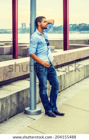 Man Thinking Outside. Wearing a light blue shirt,  blue jeans, leather shoes,  sunglasses hanging on shirt, a young guy with a little beard, mustache is leaning on a pole, sad, frowned, thinking