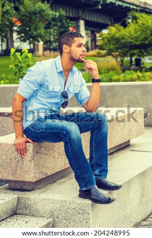 Man Thinking Outside. Wearing shirt, blue jeans, leather shoes, sunglasses hanging on shirt, hand touching his chin, a young guy with a little beard, mustache is sitting on concrete bench, thinking.