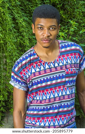 Portrait of Young Black Man. Wearing a short sleeve, collarless, colorful pattern shirt, a young handsome guy is standing by green leaves on rocks, seriously looking at you.
