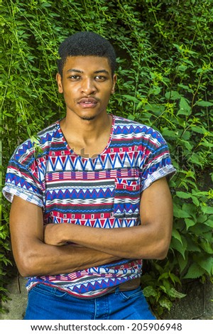 Portrait of Young Black Man. Wearing a short sleeve, collarless, colorful pattern shirt, a young handsome guy is standing by green leaves on rocks, crossing arms,  charmingly looking at you.