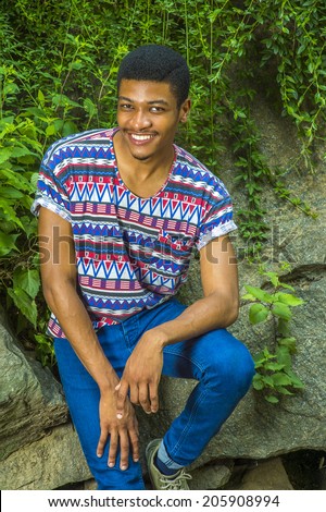 Portrait of Young Black Man. Wearing a short sleeve, collarless, colorful pattern shirt, a young handsome guy is sitting against rocks with green leaves, smiling, charmingly looking at you.