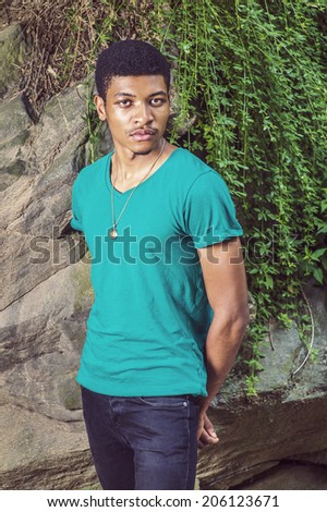 Portrait of Young Black Man. Wearing a green, short sleeve, V-neck T shirt, necklace, black pants, a young handsome guy is standing by rocks with green leaves, hands in the back, looking at you.