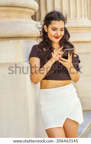 Girl Texting Outside. Dressing in black, short sleeve top, white short wrap skirt, a young lady with long curly hair is checking messages on her mobile phone, fingers touching the screen