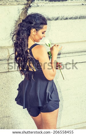 Girl Missing You. Dressing in black, sleeveless top, short pants, a young pretty lady is standing by an old fashion wall, holding a white rose, looking down, thinking.