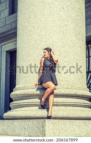 College Student. Dressing in black, sleeveless top, short pants, high heels, a young pretty lady with long curly hair is standing against a column outside an office, relaxing, thinking