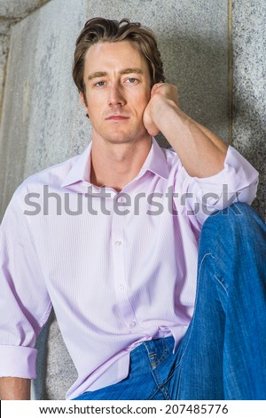 Man Thinking Outside. Wearing a light pink, long sleeve shirt, blue jeans, a young handsome guy is casually sitting against a concrete wall, thinking