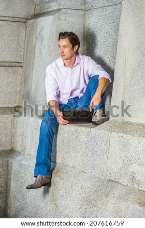 Man Working Outside. Wearing a light pink, long sleeve shirt, blue jeans, leather shoes, a young businessman is casually sitting against a concrete wall, working on a laptop computer, thinking.