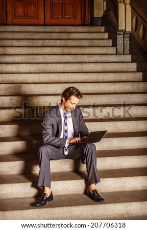 Businessman Working Outside. Dressing formally in gray suit, white under shirt, patterned neck tie, a young businessman is sitting on steps outside an office, working on laptop computer.