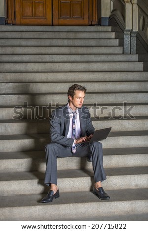 Businessman Working Outside. Dressing formally in gray suit, white under shirt, patterned neck tie, a young businessman is sitting on steps outside an office, working on laptop computer.