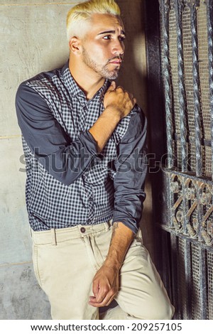 Unhappy Man. Wearing a black patterned shirt, yellow pants, a young guy with beard, yellow hair is leaning against the wall outside a metal gate, a hand on the shoulder, frowned, looking away.