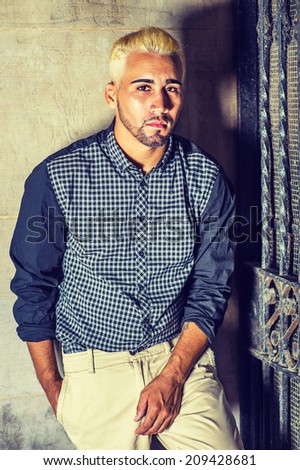Wearing a black patterned shirt, yellow pants, a young man with beard, yellow hair is leaning against the wall outside a metal gate, seriously looking at you.