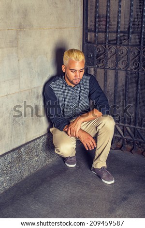 Unhappy Man Thinking on the Corner. Wearing a black patterned shirt, yellow pants, casual shoes, a young guy is squatting against the wall outside a metal gate, looking down, sad, thinking.