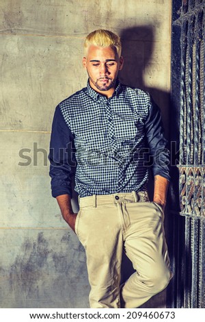 Wearing a black patterned shirt, yellow pants, a young man with beard, yellow hair is leaning against the wall outside a metal gate, hands in pockets, looking down, sad, thinking.