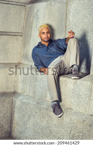Young Man Relaxing Outside. Wearing a blue shirt, gray pants, casual shoes, a young guy with beard, yellow hair is sitting down on a concrete wall, looking at you.