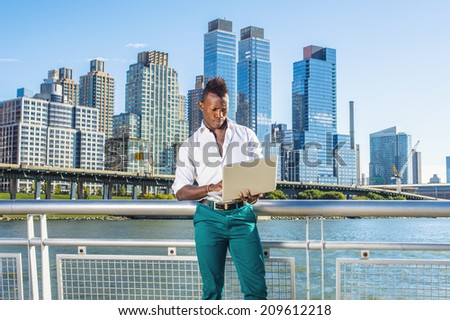 Man Working Outside. Dressing in a white shirt, green pants,  a young black guy with mohawk hair is standing in the front of high buildings, looking down, working on a computer.