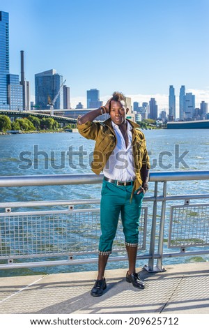 Casual Urban Man Fashion. Dressing in a yellow woolen jacket,  green pants, black leather shoes,  a young black guy with mohawk hair is standing by a river, confidently looking forward.