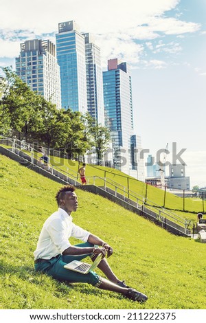 Man Working Outside. Dressing in a white shirt, green pants, black leather shoes,  a young black guy with mohawk hair is sitting on a green hill in a big city, working on a laptop computer.