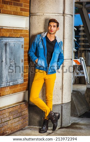 Man Thinking Outside. Dressing in a blue jacket with hood, black underwear, yellow pants, leather boot shoes, hands in pockets, a young guy is standing by a column on the corner, looking up, thinking.