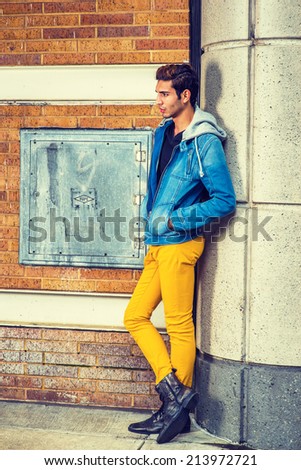 A young guy is standing by a column on the corner, relaxing, thinking wearing a blue jacket with hood, black underwear, yellow pants, leather boot shoes, hands in pockets.