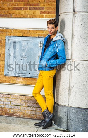 A young guy is standing by a column on the corner, relaxing, thinking wearing a blue jacket with hood, black underwear, yellow pants, leather boot shoes, hands in pockets.