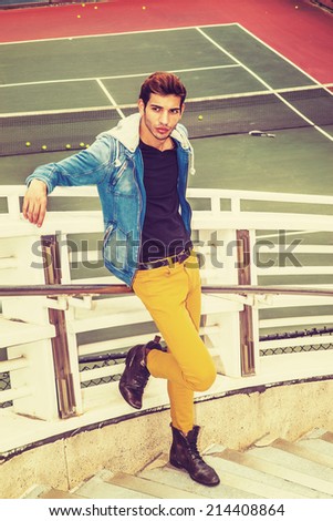 Man Casual Fashion. Dressing in blue jacket with hood, yellow pants, leather boot shoes, a young handsome guy is standing by a tennis court, arms resting on railings, relaxing, waiting, thinking.