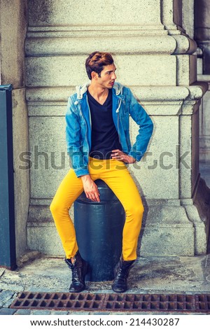 Man Fashion. Dressing in a blue jacket with hood, black underwear, yellow pants, leather boot shoes, a young handsome guy is sitting on a metal stake on the corner, looking around, waiting for you.