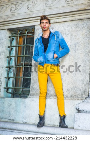 Man Casual Fashion. Dressing in a blue jacket with hood, black underwear, yellow pants, leather boot shoes, hands in pockets, a young handsome guy is standing by a window, relaxing, waiting for you.